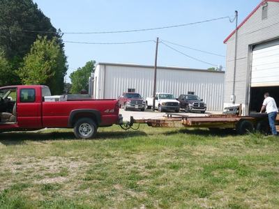 Red pickup truck hitched to flatbed trailer with man standing at trailer near garage
