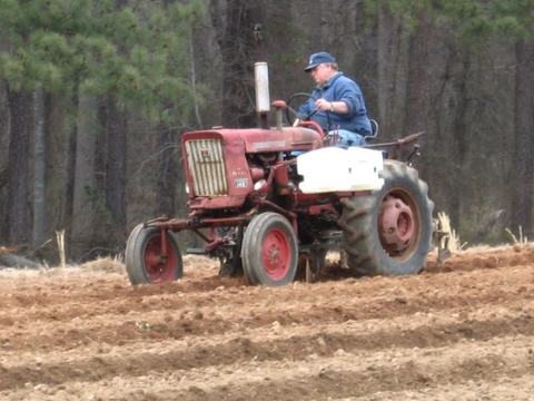 Person driving a red tractor tilling parallel furrows in a field with trees behind