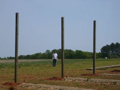 Man standing in field among three upright wooden posts and prepared planting rows