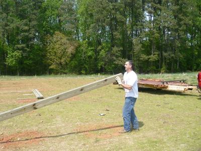 Man wearing gloves carries a long wooden beam across a grassy field near trees.