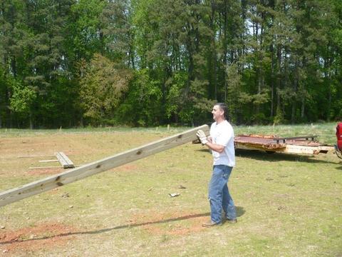 Man wearing gloves carries a long wooden beam across a grassy field near trees.