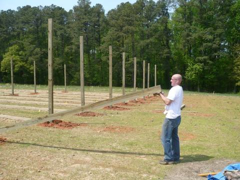 Man carrying a long wooden plank near rows of upright fence posts in a field