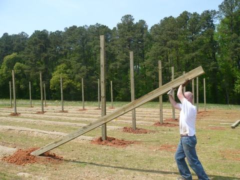 Man lifting a long wooden beam toward upright posts on a cleared field