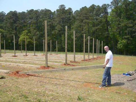 Man carrying a long wooden beam at a construction site with rows of upright posts