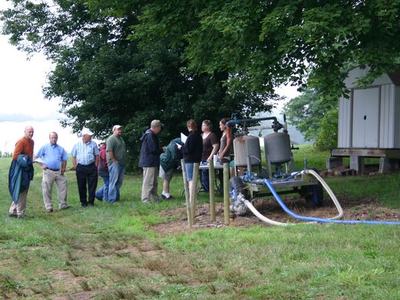People gathered by a portable water pump with hoses near a shed under trees