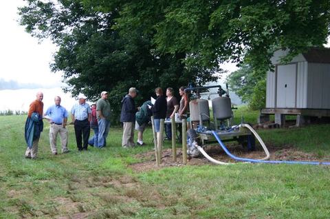 People gathered by a portable water pump with hoses near a shed under trees