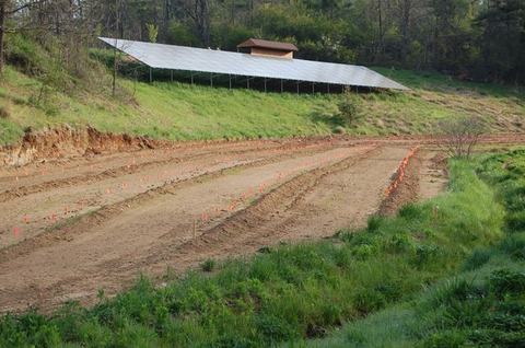 Plowed field with orange marker flags below a hillside solar panel array and small shed