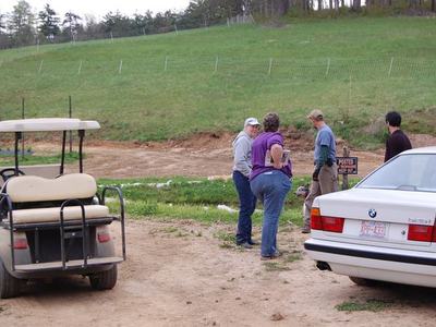 Four people standing and talking near a parked golf cart and car on dirt by a grassy hill