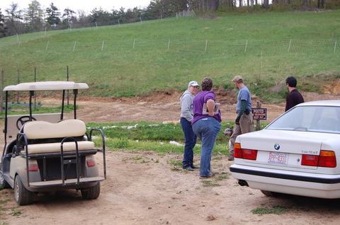 Four people standing and talking near a parked golf cart and car on dirt by a grassy hill