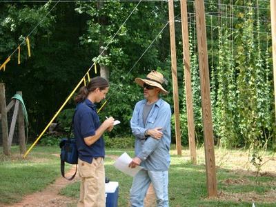 Two men in a hop field talking; one takes notes while hop vines climb trellis