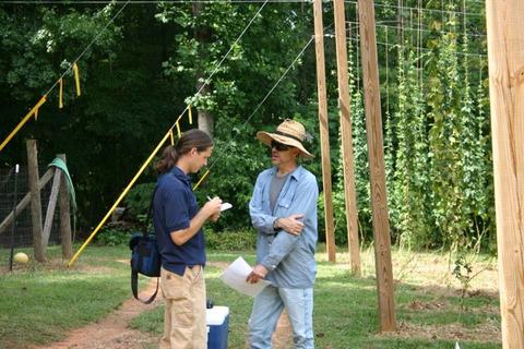 Two men in a hop field talking; one takes notes while hop vines climb trellis