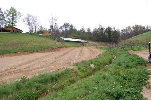 Graded dirt road curving through grassy hillside toward a low barn and houses in distance