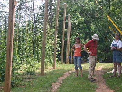Three people standing beside tall wooden posts with climbing vines in a grassy wooded area