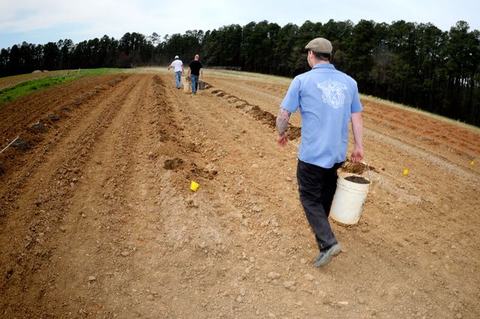 Three people walking across a plowed field, one carrying a white bucket