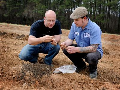 Two men kneeling on bare soil examining small plant roots or tubers beside a plastic bag.