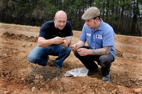 Two men kneeling on bare soil examining small plant roots or tubers beside a plastic bag.