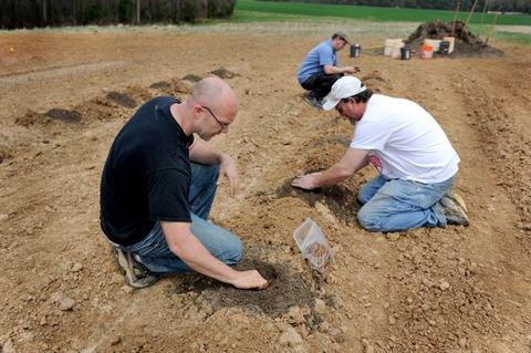 Three men kneeling in a field planting seeds into prepared soil rows