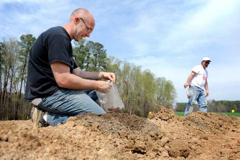 Man kneeling on field placing soil into a plastic bag; another man walking in background