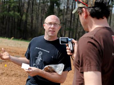 Bald man holding plastic bag of soil and paper gestures while another man films him outdoors