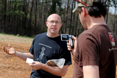 Bald man holding plastic bag of soil and paper gestures while another man films him outdoors