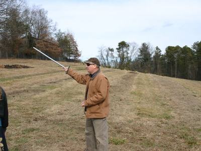 Man in brown jacket and cap pointing a long pole across a grassy field