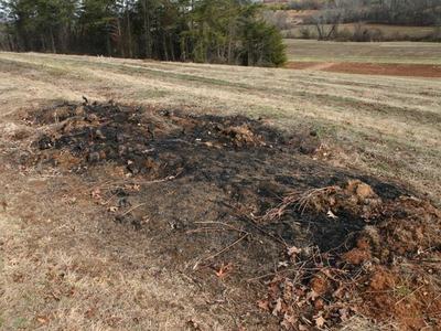 Burned patch of grass and soil in a mowed field with trees in background