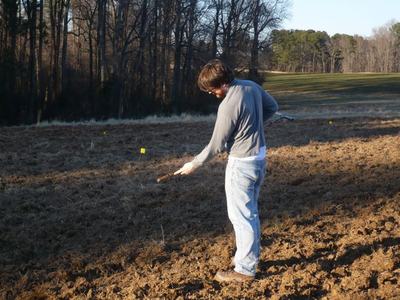 Person in gray shirt and jeans using a stick in a plowed field with yellow markers