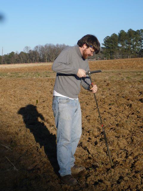 Man wearing sweater inserting a soil probe into a plowed field