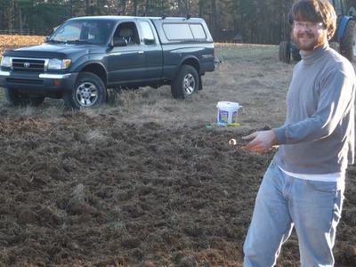 Man standing in a plowed field holding a small object, pickup truck parked behind