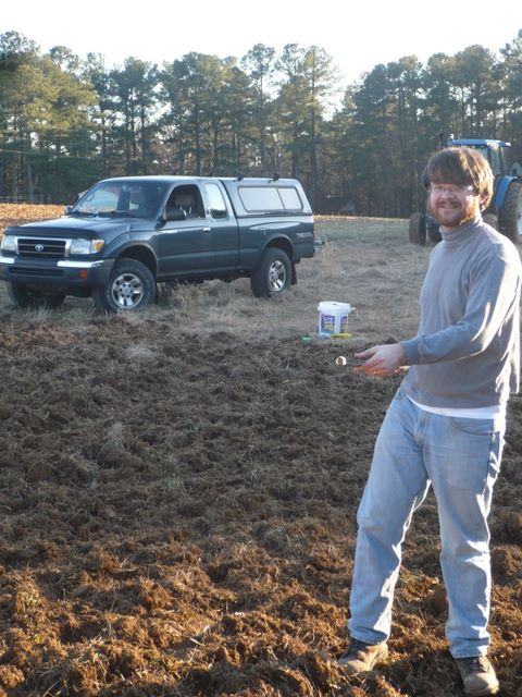 Man standing in a plowed field holding a small object, pickup truck parked behind