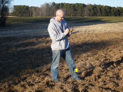Man in hoodie standing in a grassy field using a handheld GPS; yellow marker at his foot