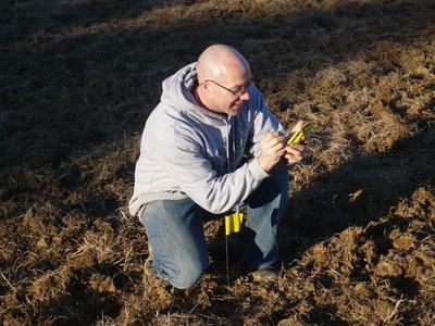 Person kneeling in dry field holding and inspecting a small yellow handheld device