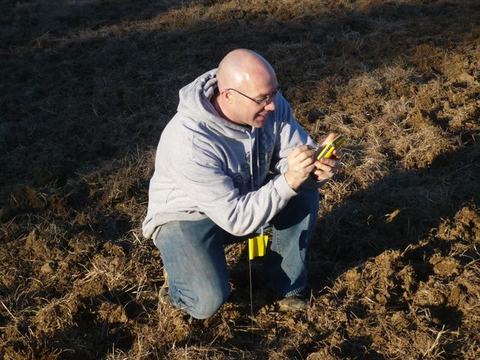 Person kneeling in dry field holding and inspecting a small yellow handheld device