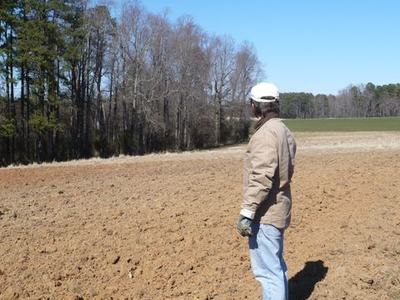 Person wearing cap and jacket standing on a plowed field looking toward tree line