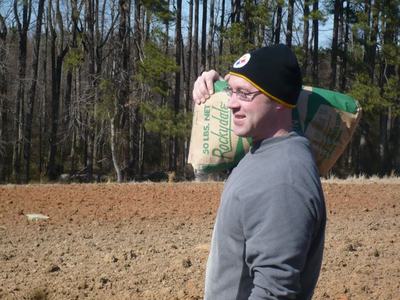 Man wearing beanie carries a bag labeled "50 LBS. NET Rockdale" over shoulder in a plowed field