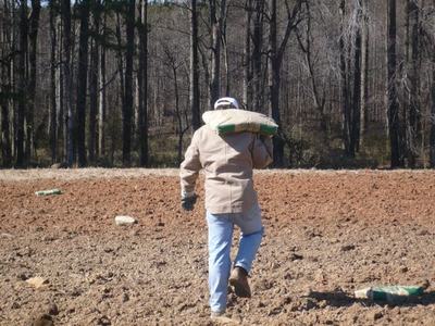 Person carrying sack over shoulder walking across plowed field toward woods