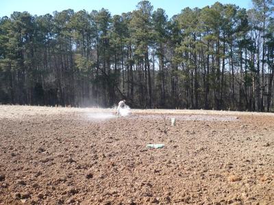 Person riding an ATV across a plowed field, kicking up a cloud of dust, trees in background