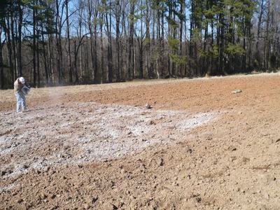 Person scattering white lime or fertilizer on a freshly plowed field near a treeline