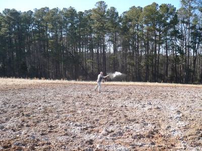 Man scattering white powder across plowed field with tree line in background