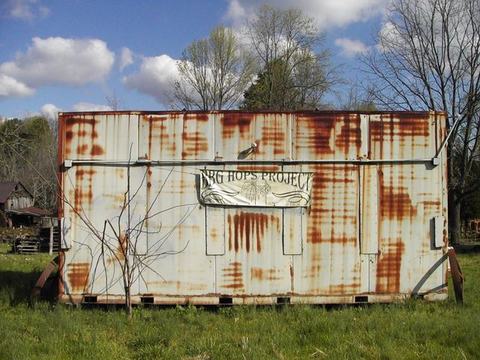 Rusty white metal shipping container with "BIG HOPS PROJECT" banner in a grassy field