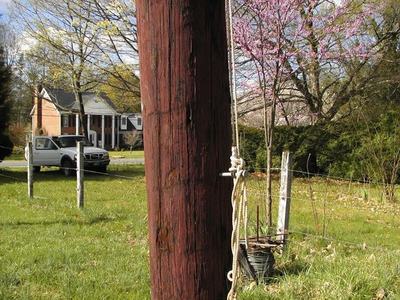Wooden utility pole with coiled rope and pulley beside grassy yard; house and truck in background