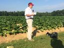 Man in red visor holding microphone and papers standing at edge of a green crop field