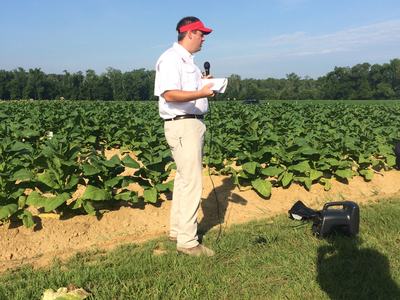 Man in red visor holding microphone and papers standing at edge of a green crop field