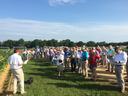 Man speaking to group of people at outdoor agricultural field day, attendees with papers
