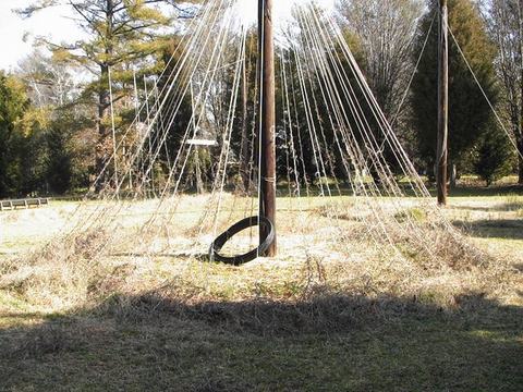 Central wooden pole with many radiating ropes, small hanging swing and tire at base in grassy field