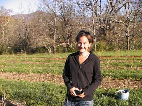 Woman standing in grassy field holding a small object; bucket with harvested items nearby