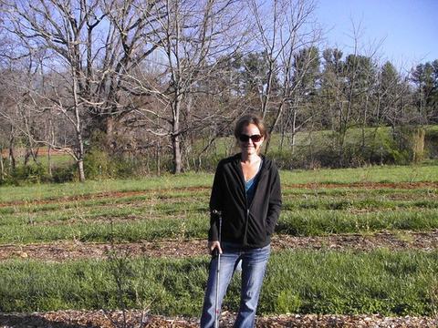 Woman in sunglasses and jacket standing in a grassy field with leafless trees