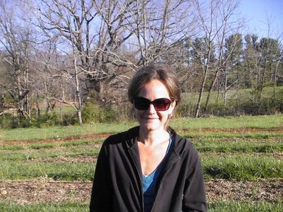 Woman wearing sunglasses and jacket standing in a grassy field with leafless trees