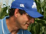 Man in blue UK baseball cap and blue polo, cigarette in mouth, standing by corn stalks.