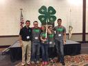 Five young people standing on a stage holding a large green 4-H clover sign, US flag behind